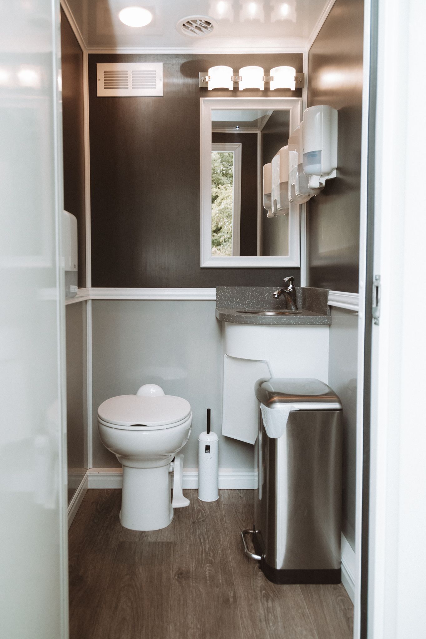 Interior of a portable restroom with a toilet, sink, mirror, and trash can. Dark and light tones — Absoloo Hire Pty Ltd In Morisset, NSW