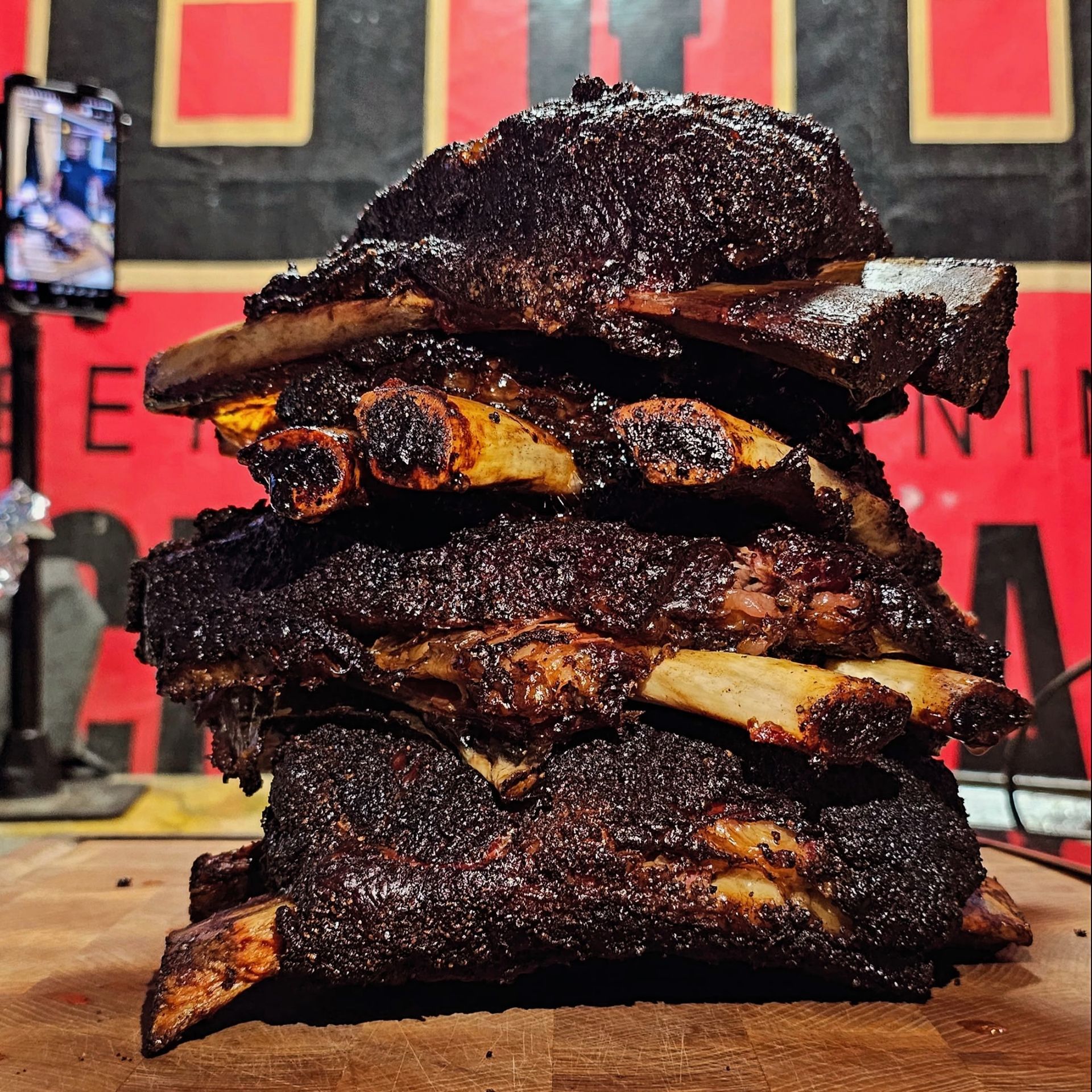 Stack of smoked beef ribs on a wooden cutting board, with a dark, crusty bark. Red and black background.