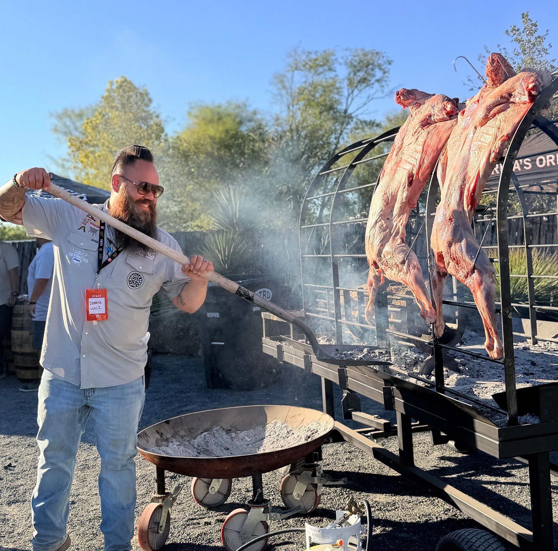B&B Team pitmaster Derrick Walz cooking meat on a spit over an open fire outdoors near a large pan.