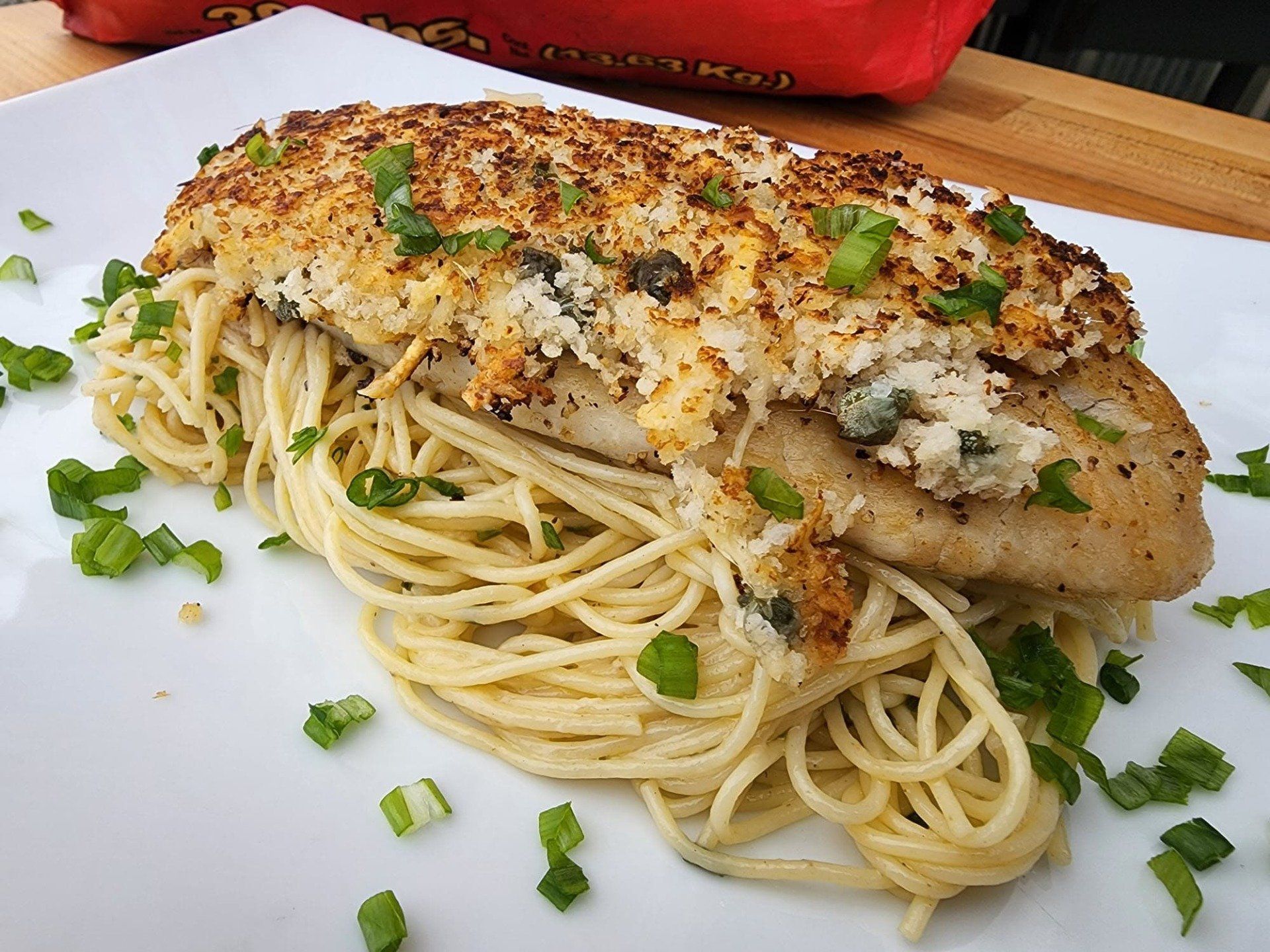 A breaded, grilled fish fillet topped with herbs sits on a bed of spaghetti on a white plate, sprinkled with green onions.