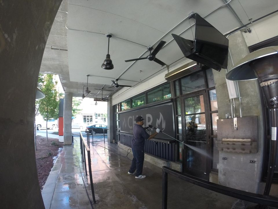 Person standing outside a storefront under an awning, washing the sidewalk with a hose.