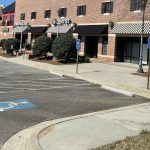 Brick storefronts with awnings and trimmed shrubs along a sidewalk and parking lot.