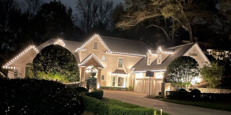 House outlined with warm white Christmas lights at night, framed by trees and a lit driveway