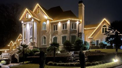 Large two-story house outlined with warm white holiday lights at night, with palm trees and landscaped front yard