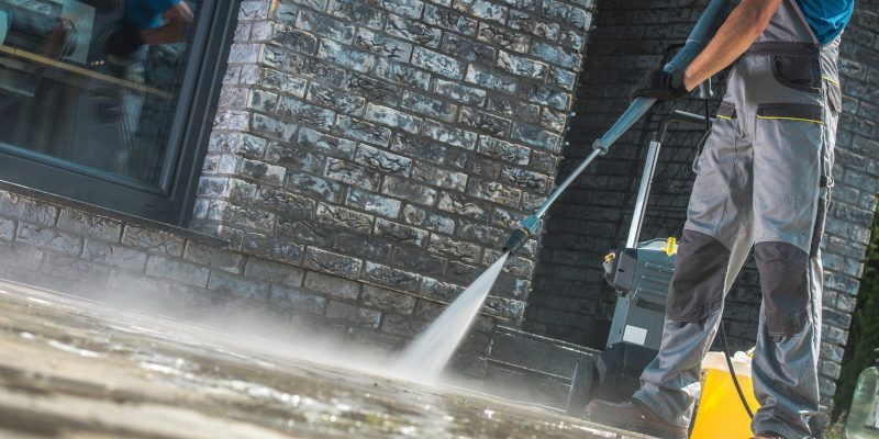 Worker pressure-washing a brick wall beside a sidewalk, with spray mist and cleaning equipment visible