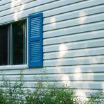 Blue shuttered window on white house siding with sunlight and greenery below