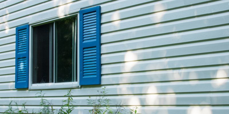 Blue shuttered window on a white siding house with sunlight and plants below
