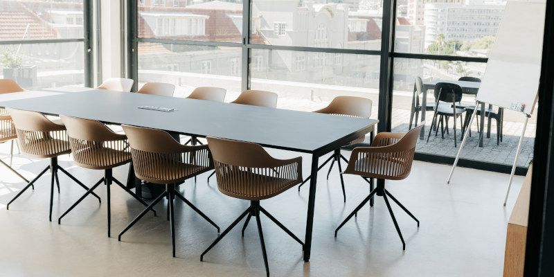 Modern conference room with a long table, woven chairs, and floor-to-ceiling windows overlooking a city.