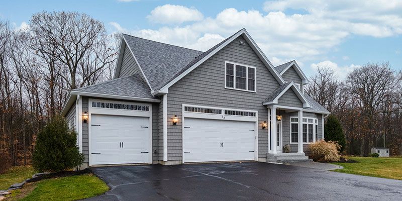 Gray two-story suburban house with white trim, three garage doors, and a front porch under a cloudy sky