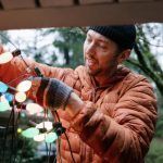 Person in a brown jacket hanging colorful string lights outdoors at dusk