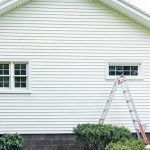 Ladder leaning against a white house beside a small window, with bushes below.