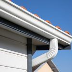 White gutter and downspout on a house roof edge with orange trim against blue sky