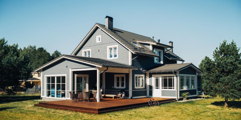 Large gray house with white trim and porch, surrounded by trees under a clear blue sky