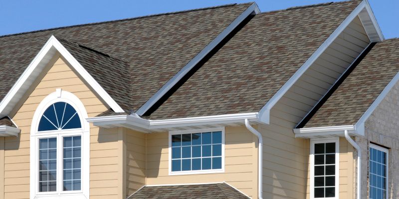 Suburban house roof with tan siding, white trim, and windows under a blue sky