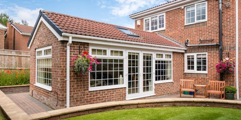 Brick house sunroom with white-framed windows, patio doors, and hanging flower baskets beside a paved driveway