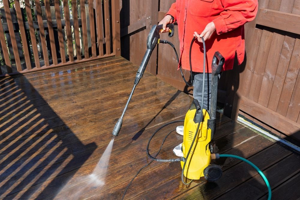 Person pressure-washing a wooden deck with a yellow cleaner on a sunny day