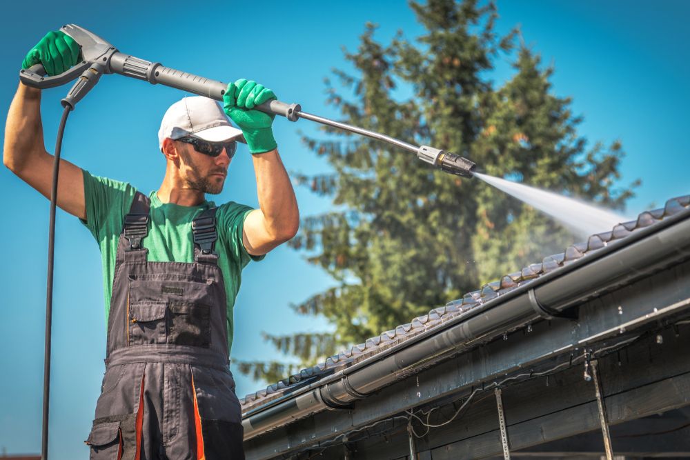 Worker spraying roof with hose from ladder, wearing green gloves and dark overalls under a blue sky