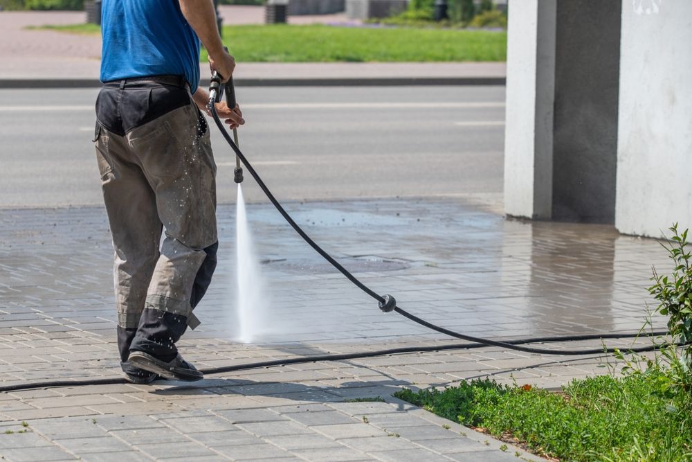 Person pressure-washing a sidewalk with a hose beside a street and building entrance