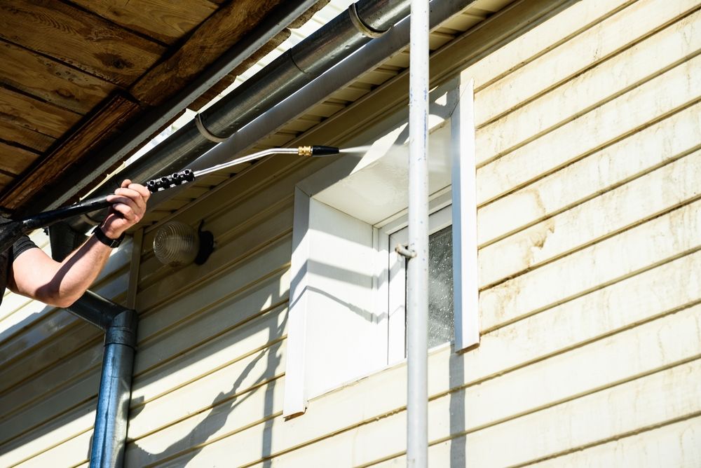 Person pressure-washing tan house siding with a long wand from a ladder