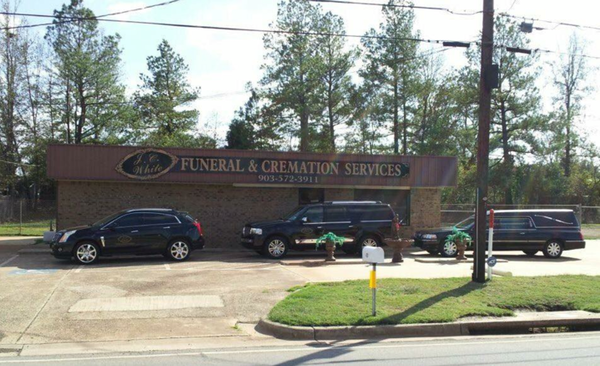 Funeral home with hearse and cars parked out front, brown building, sunny day.