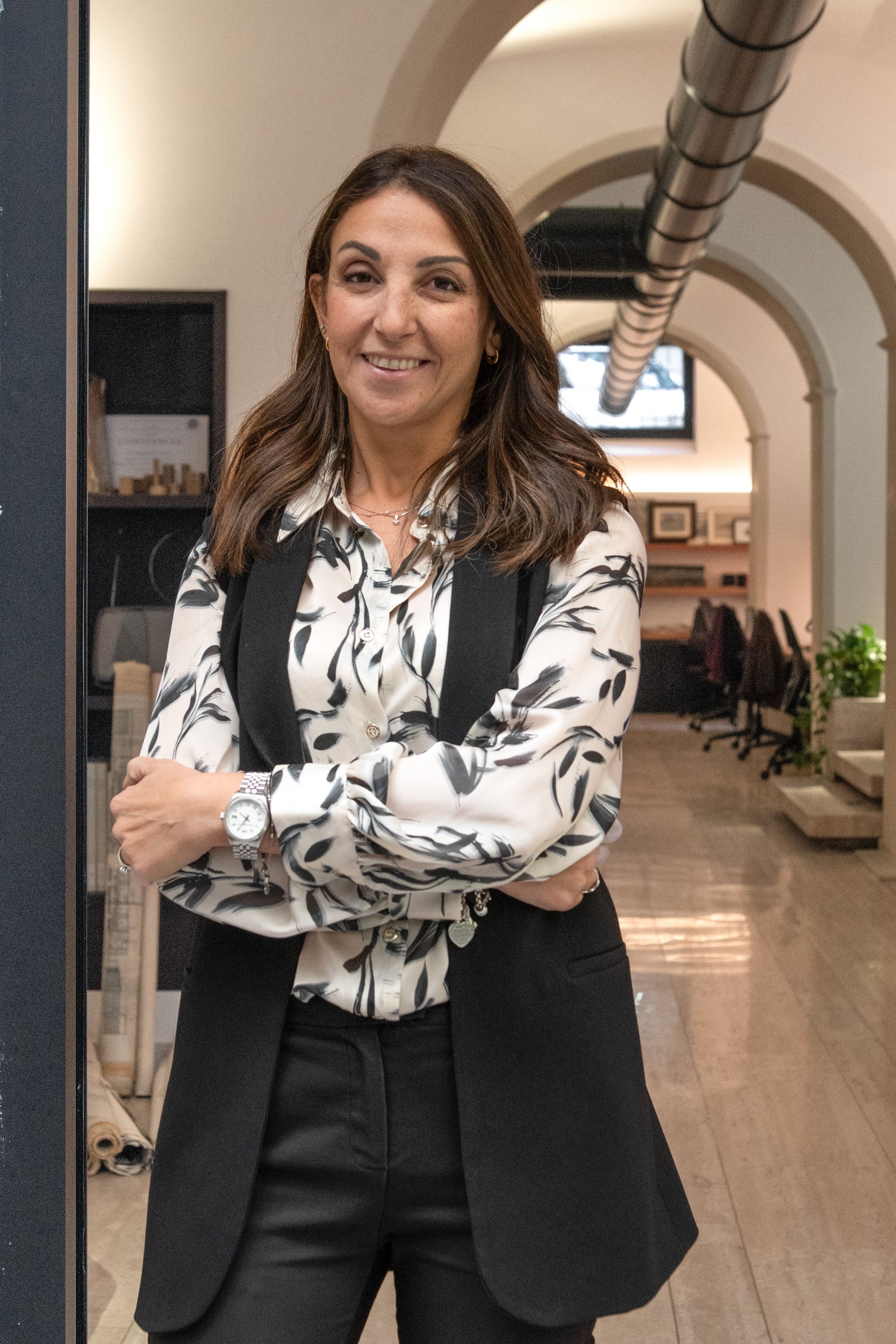 Donna in piedi in un corridoio ad arco, con una camicetta bianca a fiori e un blazer nero, sorridente a braccia conserte