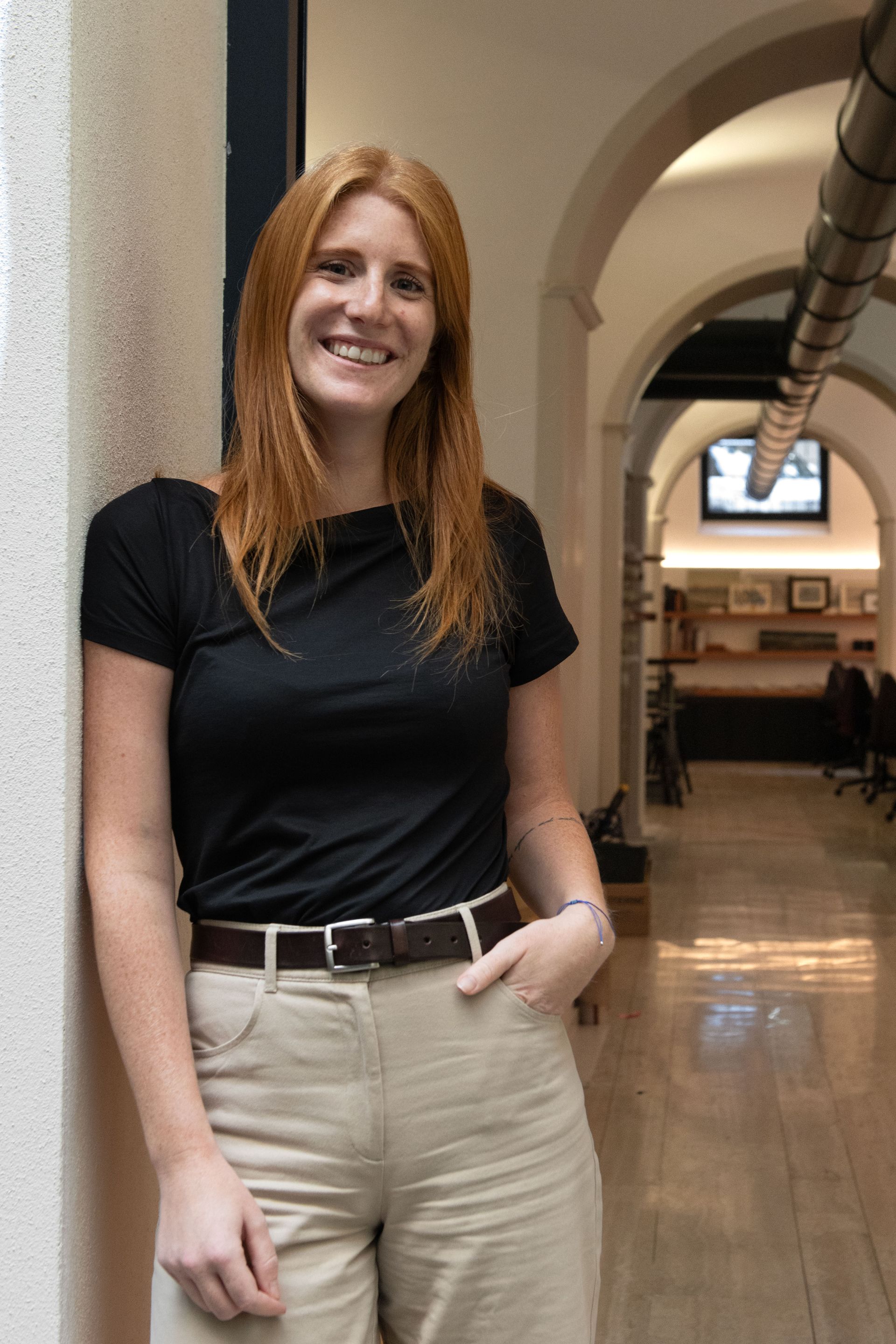 Una persona sorridente con una maglia nera e pantaloni beige è in piedi in un corridoio con soffitti a volta.