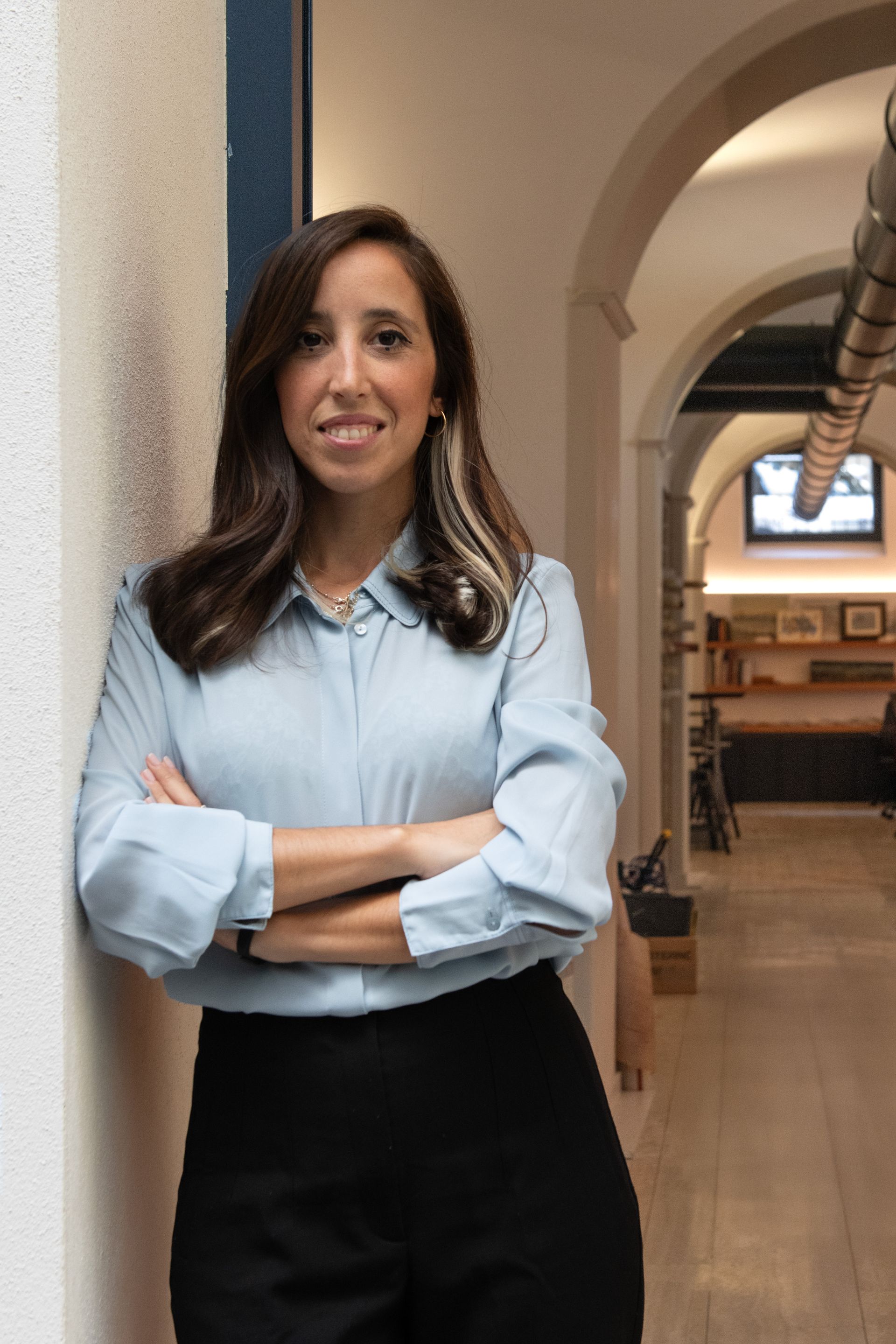 Donna con una camicia azzurra, in piedi con le braccia conserte in un corridoio, che sorride.