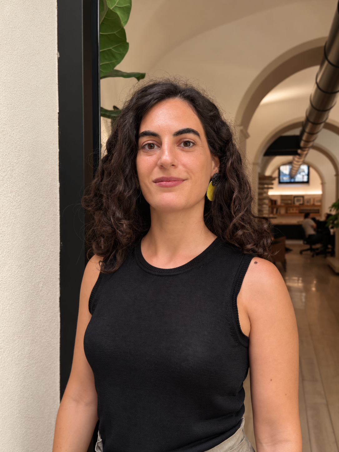 Persona con i capelli scuri e ricci, in un top nero senza maniche, in piedi in un corridoio interno ad arco.