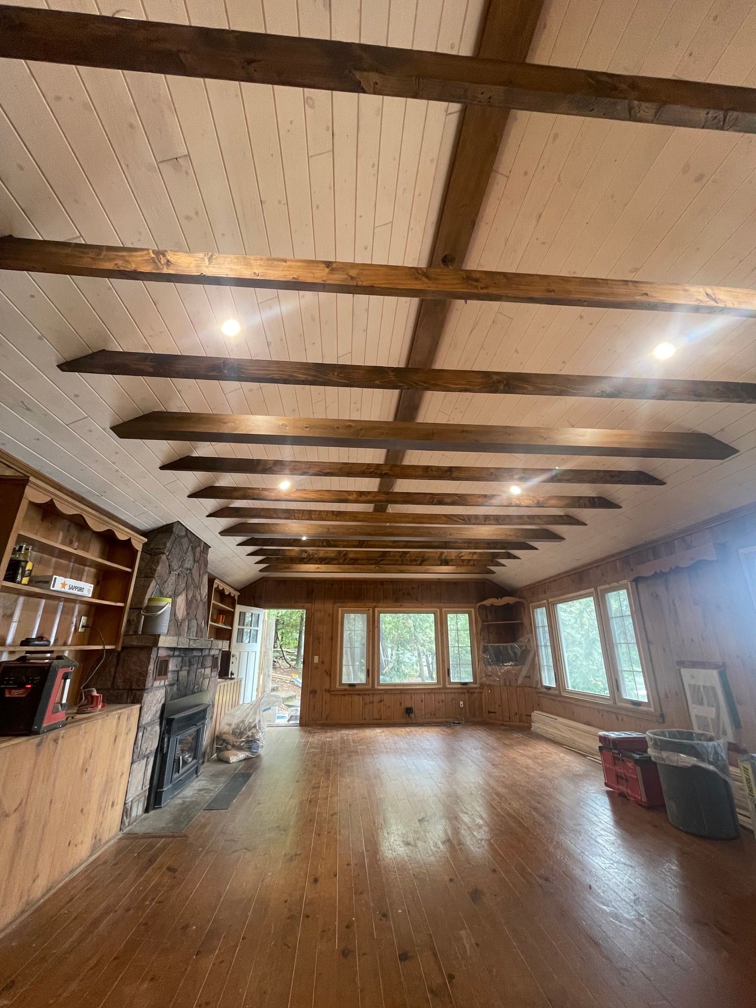 Wooden beamed ceiling in a cabin-like interior; hardwood floors, fireplace, windows, and built-in shelving.