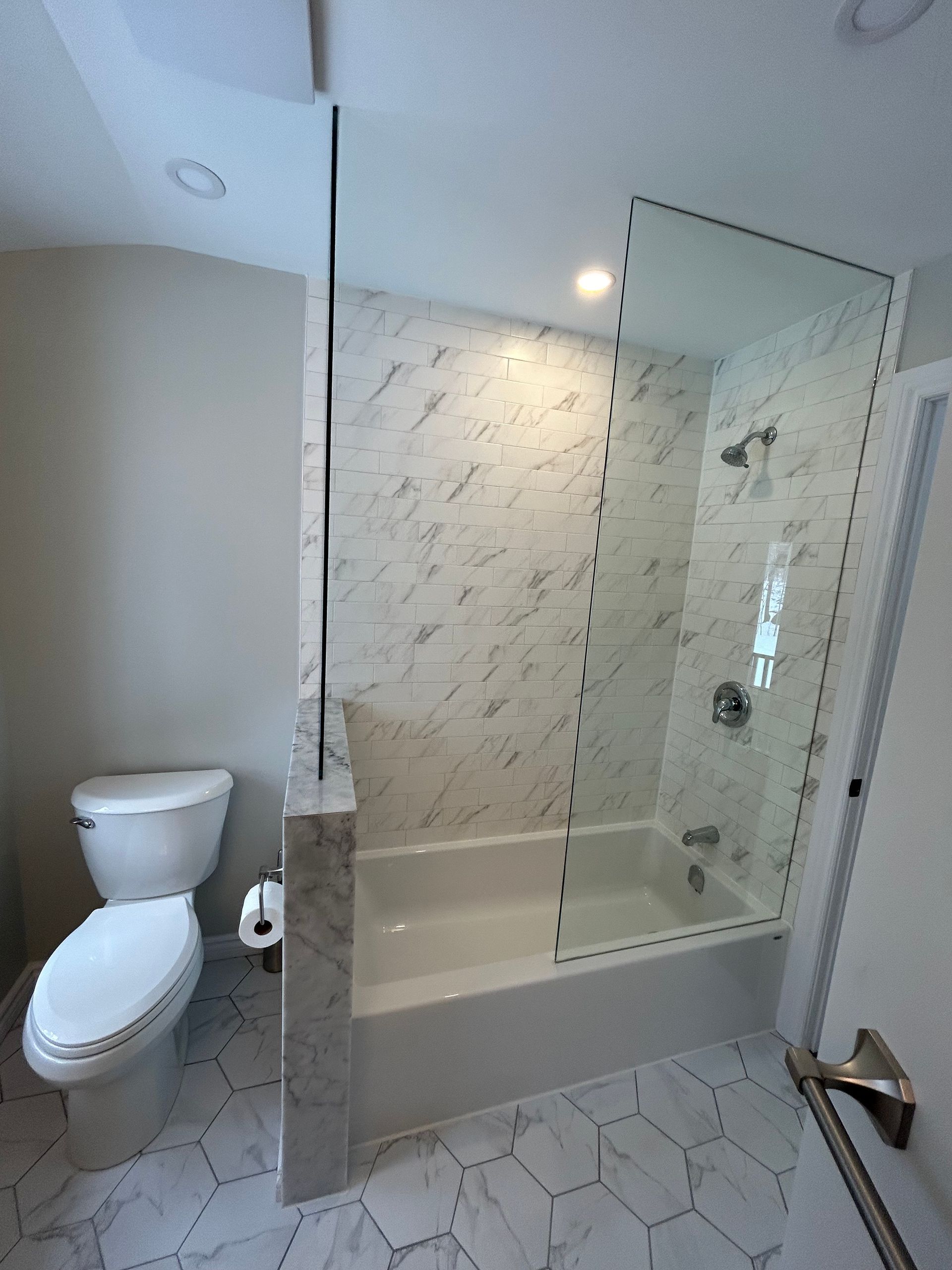 Bathroom with tub, marble-patterned tile, glass shower panel, and a toilet on a patterned floor.
