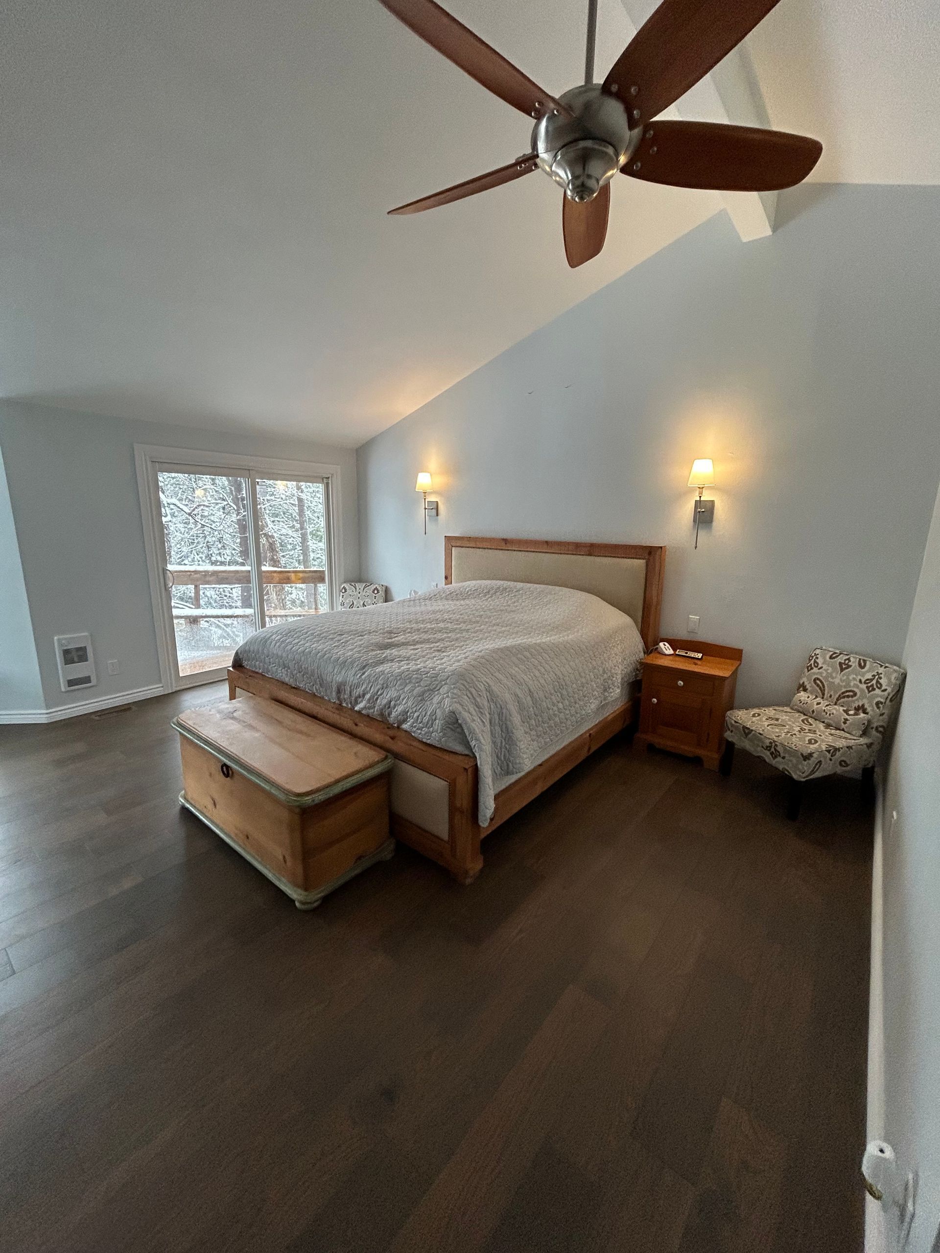 Bedroom with wooden bed, nightstand, and armchair; large windows overlook snowy trees.