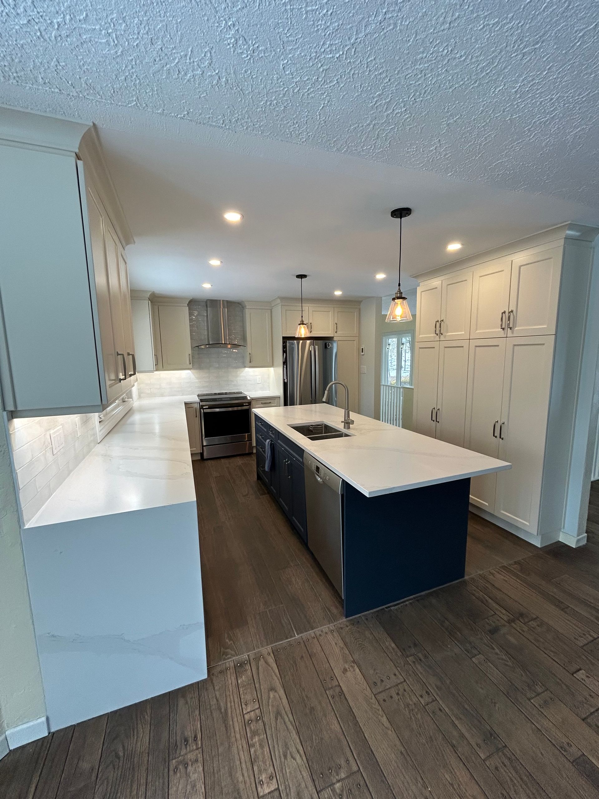 Modern kitchen with white countertops and cabinets, a navy blue island, stainless steel appliances, and wood flooring.