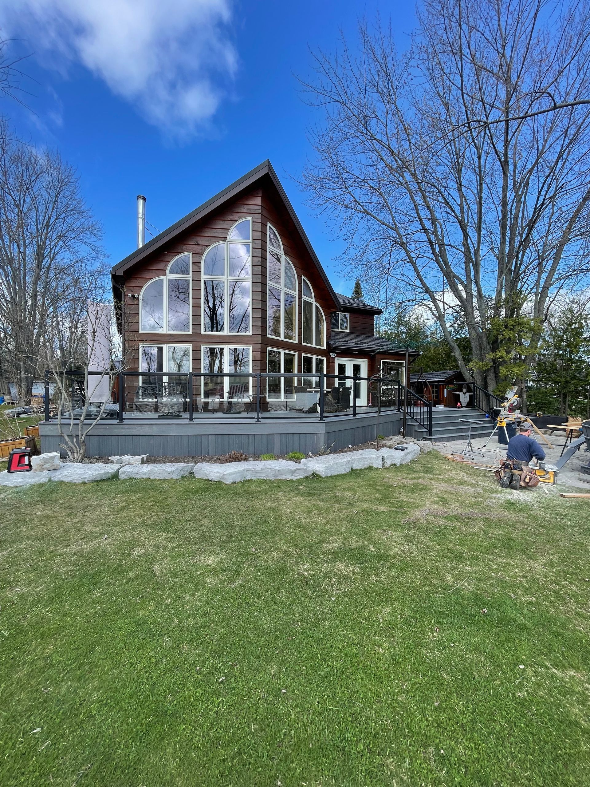 Log cabin with large windows and a wraparound deck on a grassy lawn under a blue sky.