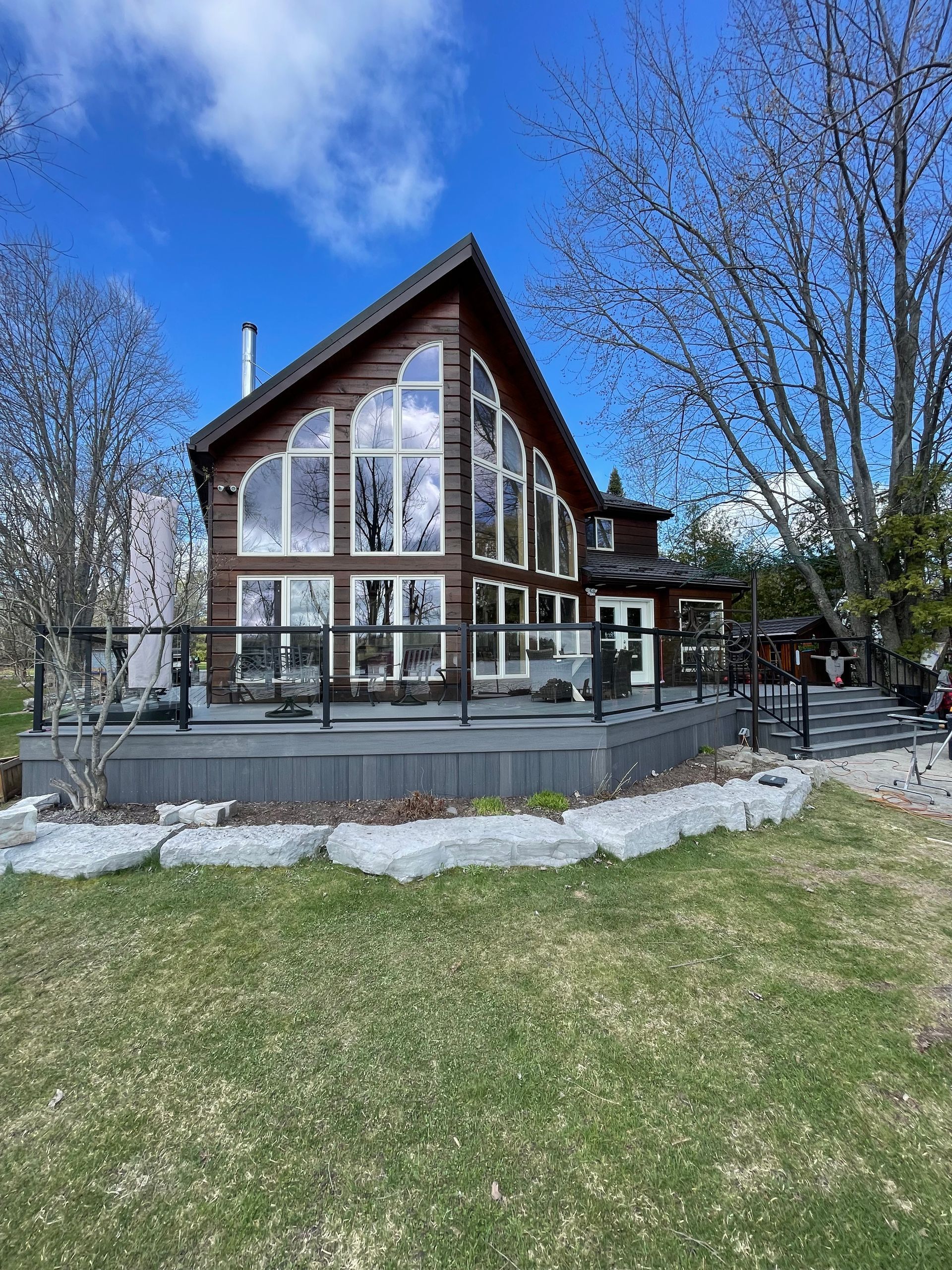 Rustic brown cabin with large windows, surrounded by a deck and grassy yard, under a blue sky.
