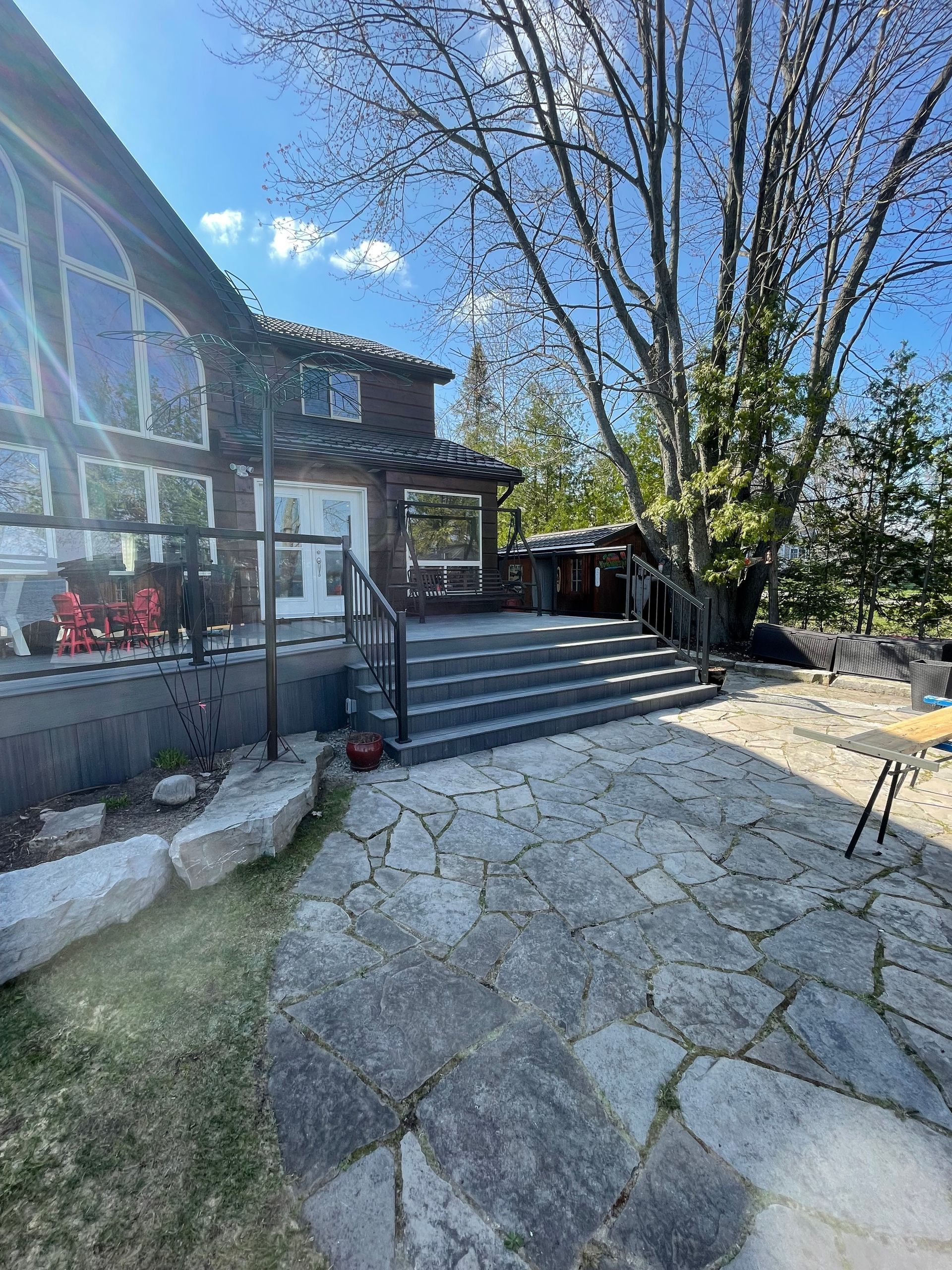 Stone patio and steps leading to a wooden deck attached to a house with large windows under a blue sky.