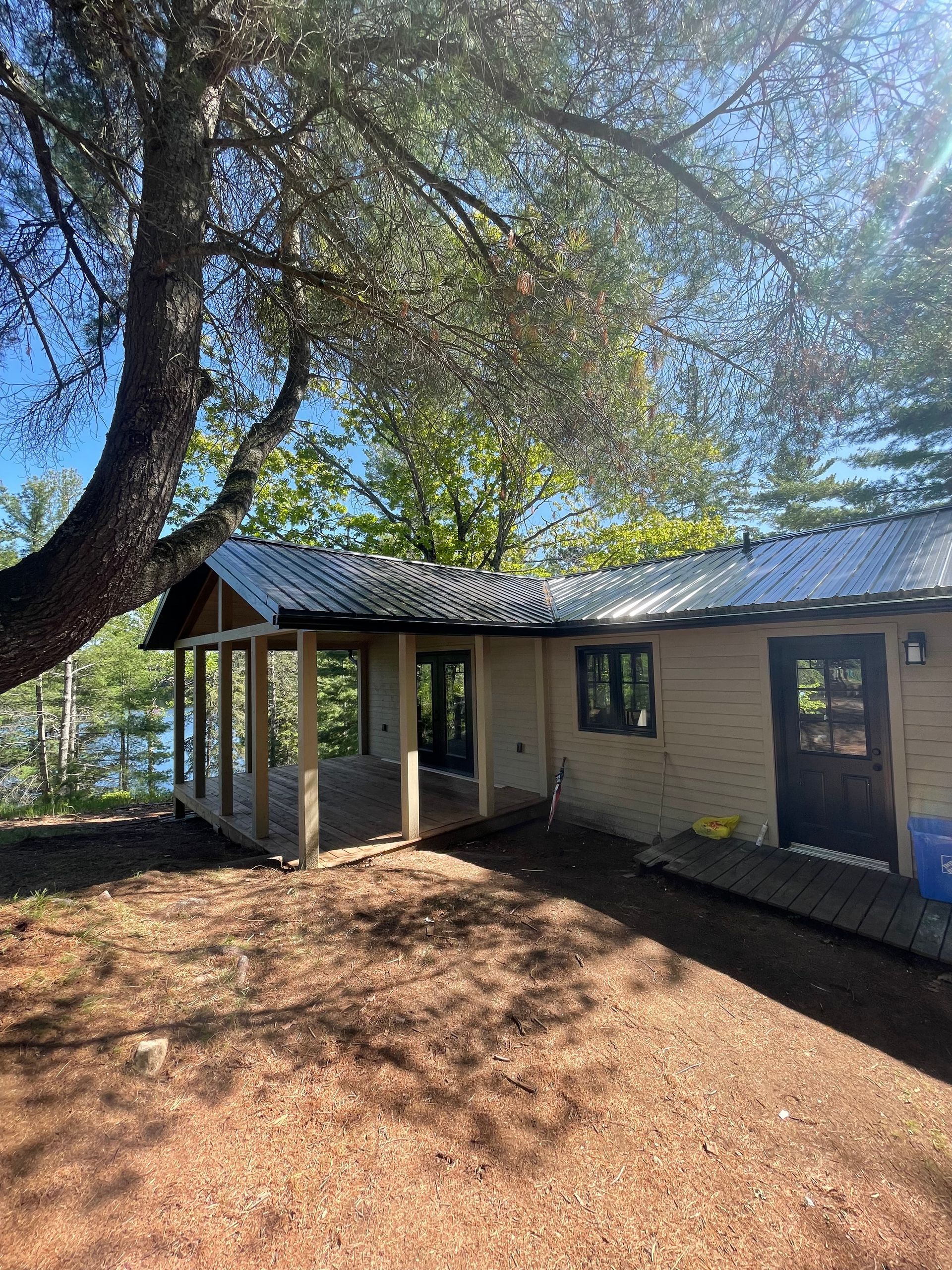 Small beige building with a porch under a tree, beside a body of water.