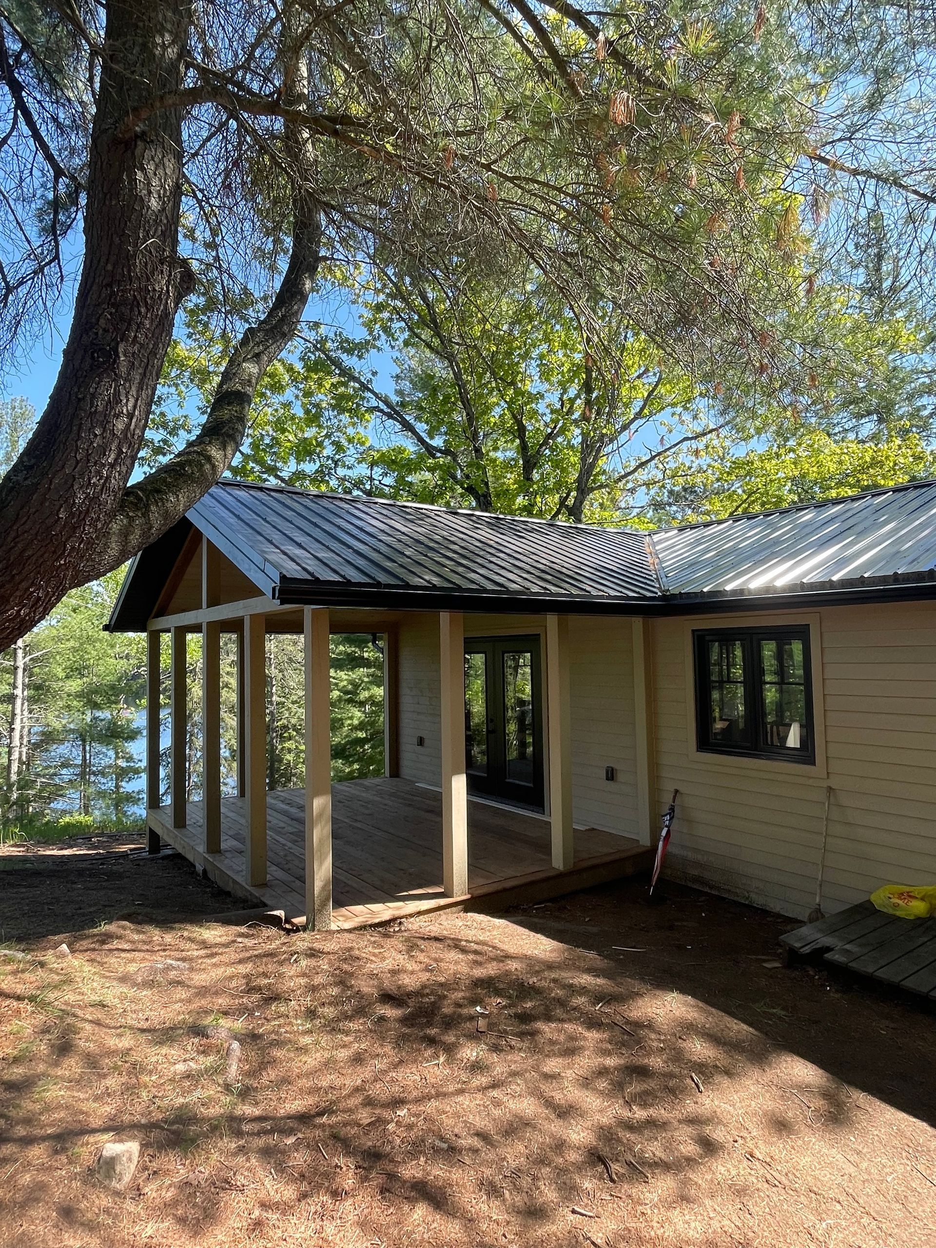 Small cabin with a covered porch, near a lake, under a tree.