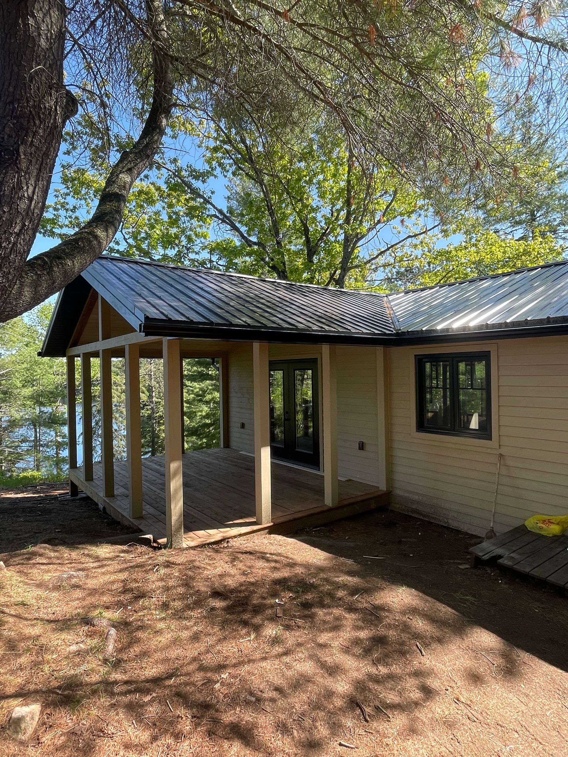 Cabin with a covered porch and metal roof, set in a wooded area with trees and a lake in the background.