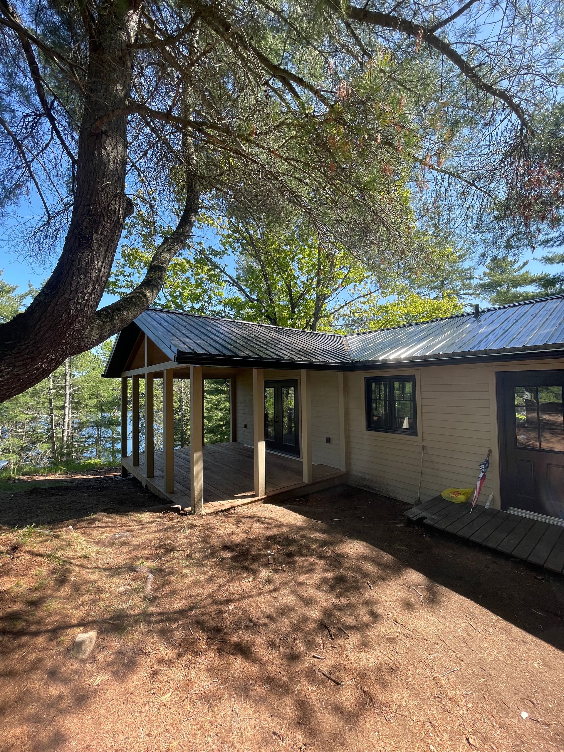Cabin with porch under tree overlooking water. Brown building with dark roof and wooden supports.