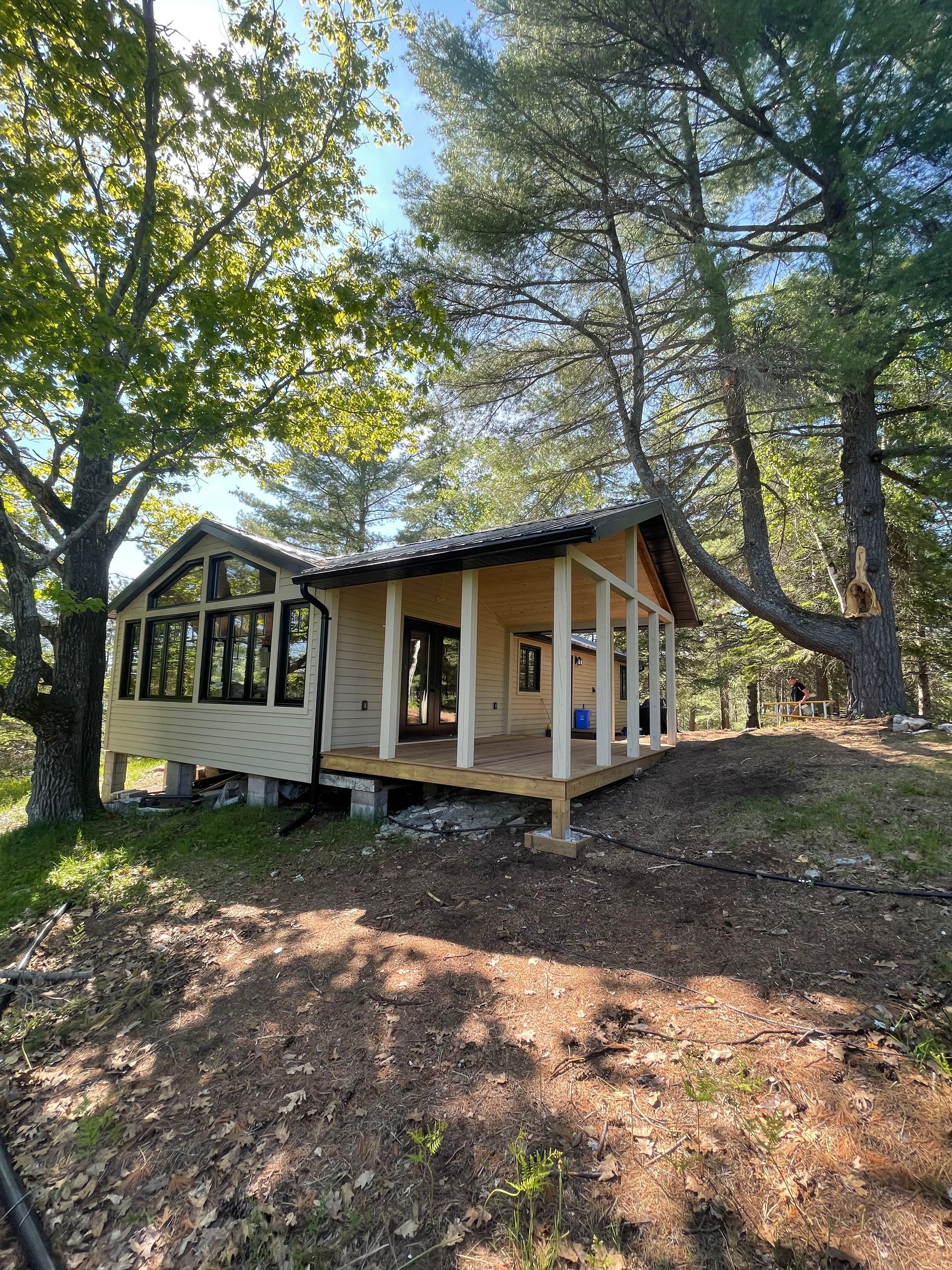 Small wooden cabin on a hillside, with a porch. Surrounded by trees, under a bright sky.