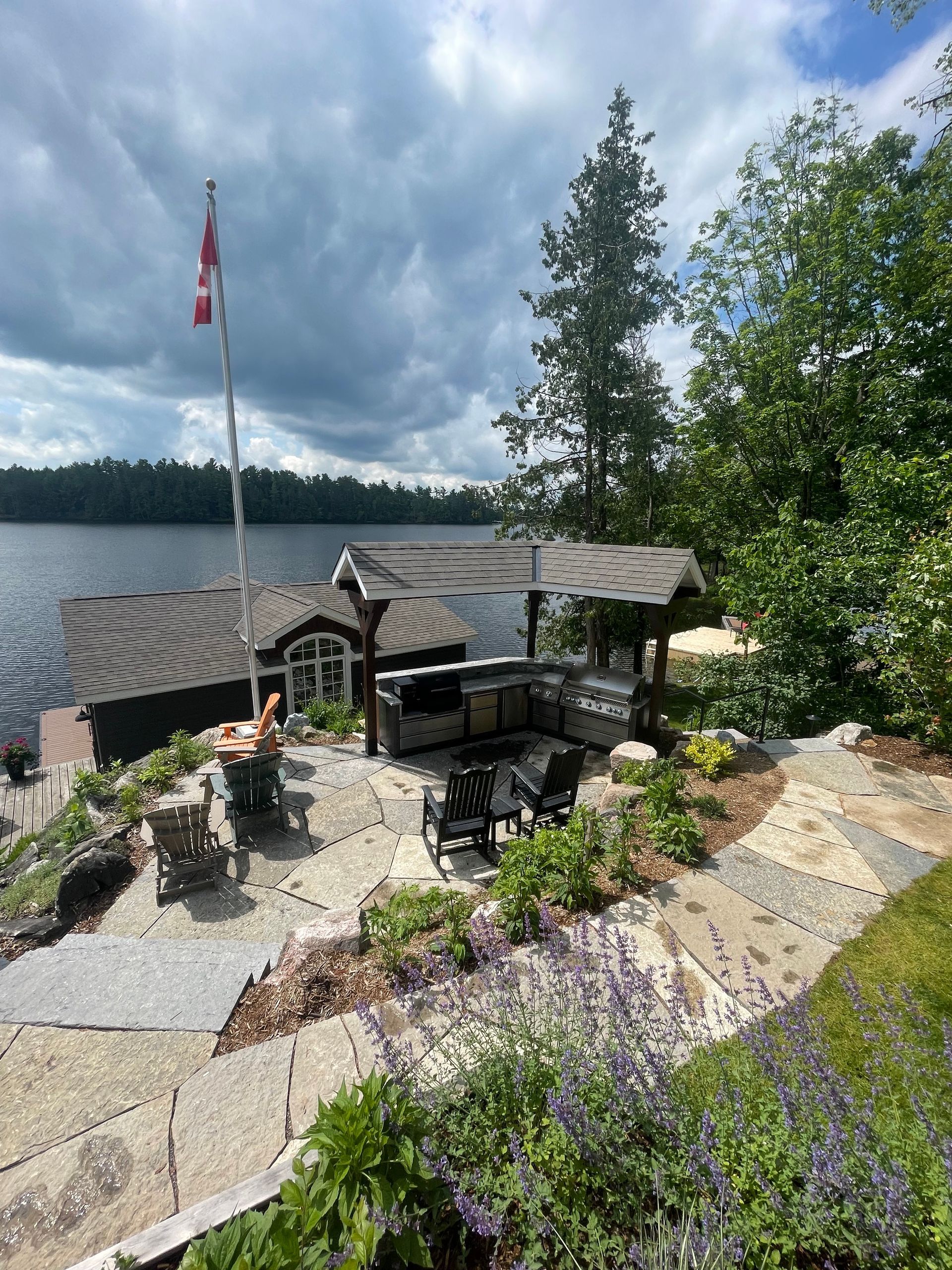Lakeside cabin with patio, outdoor kitchen, Canadian flag, and lake view under a cloudy sky.
