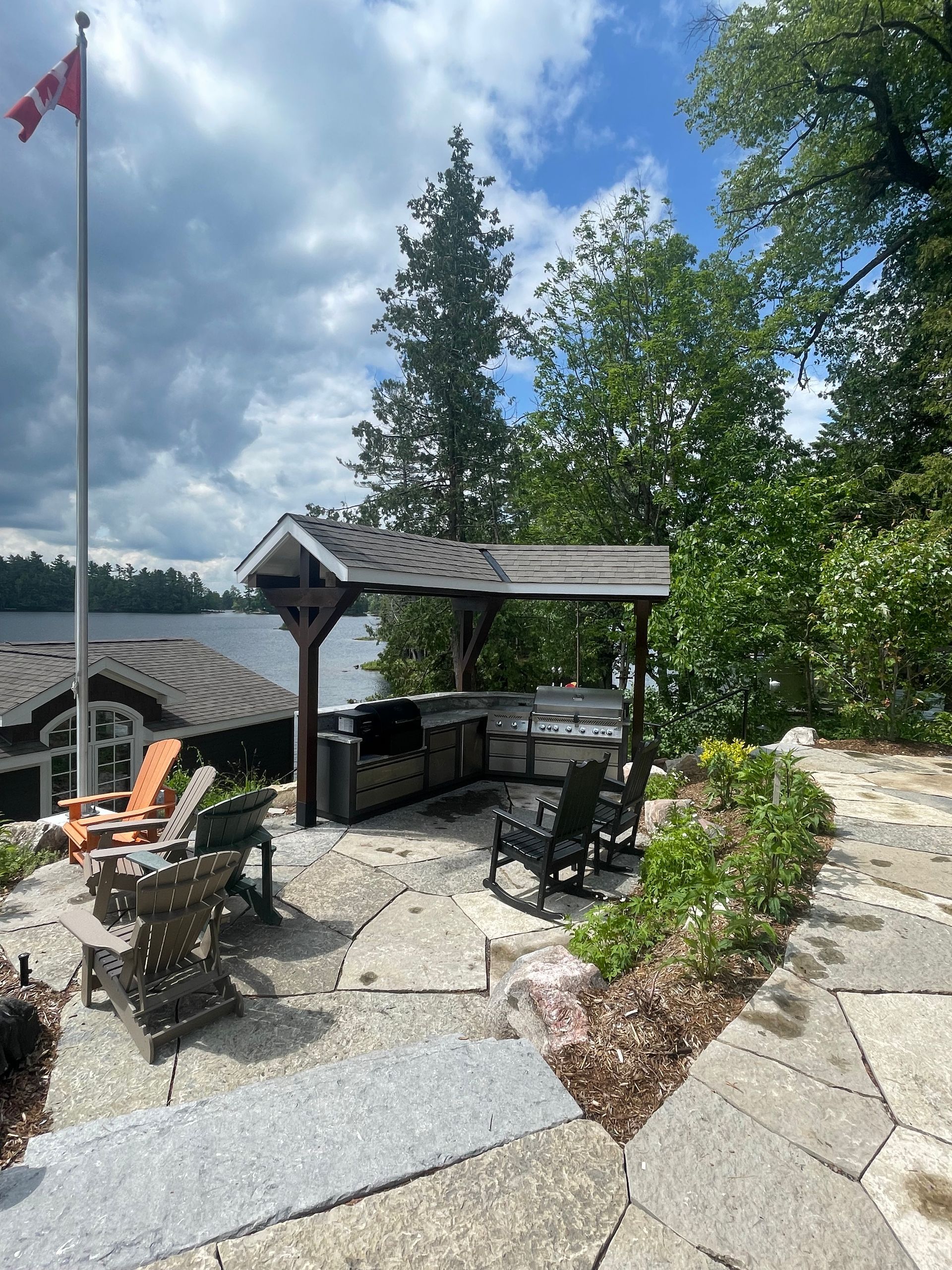 Outdoor kitchen with grilling area and seating, overlooking a lake under a cloudy sky.