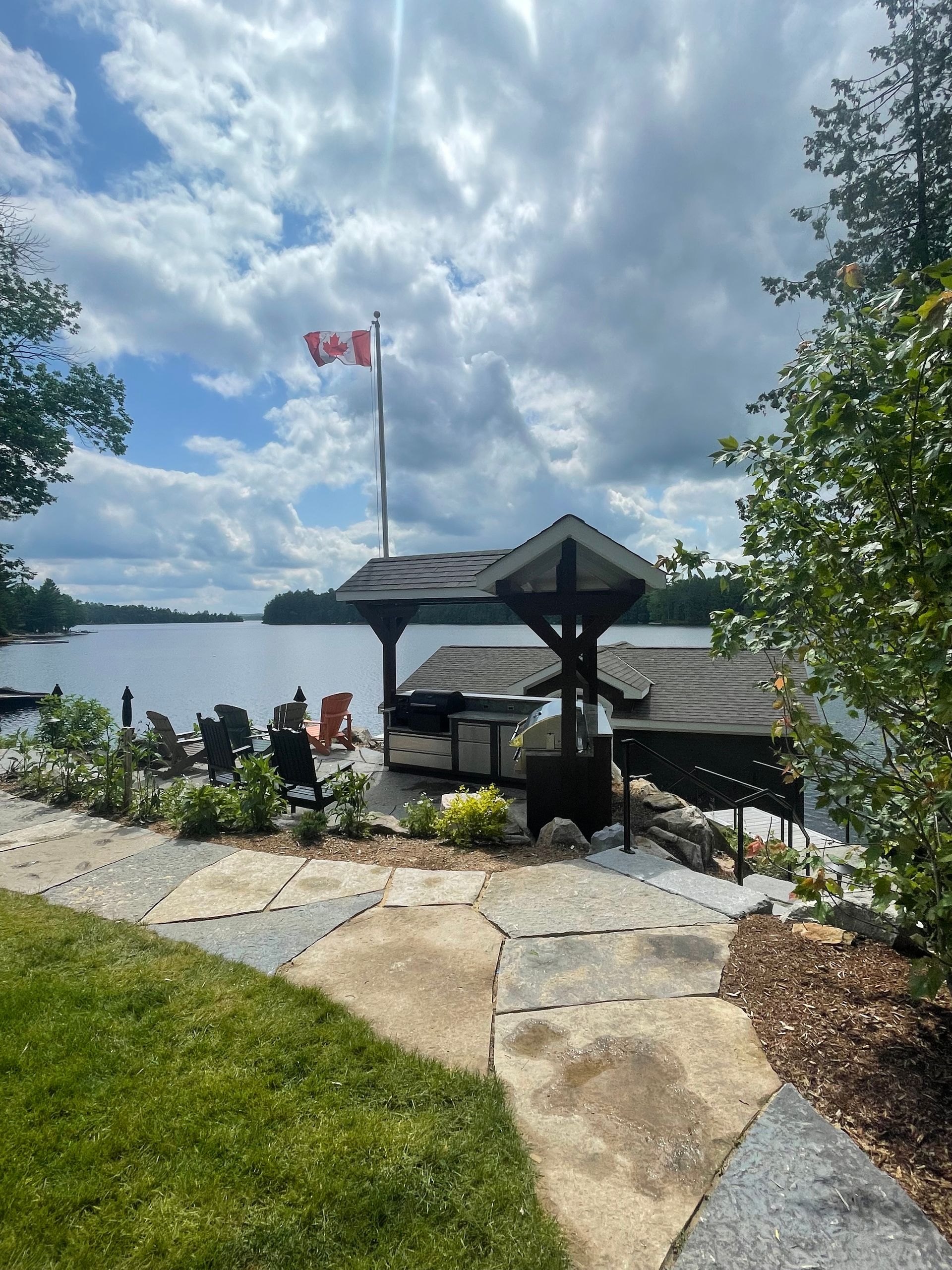 Lakeside patio with gazebo and Canadian flag. Cloudy sky, water view, stone path, green grass.