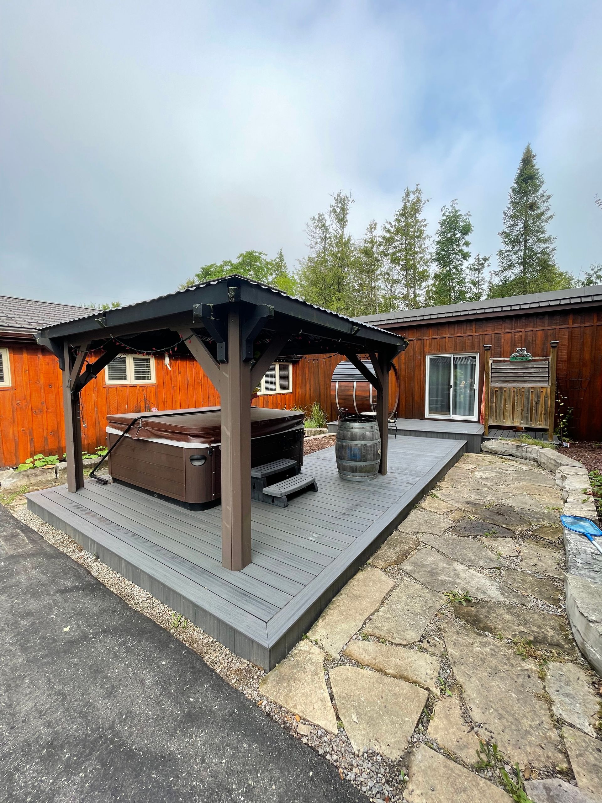 Hot tub under a dark gazebo on a gray deck next to a building with reddish-brown siding and a stone patio.