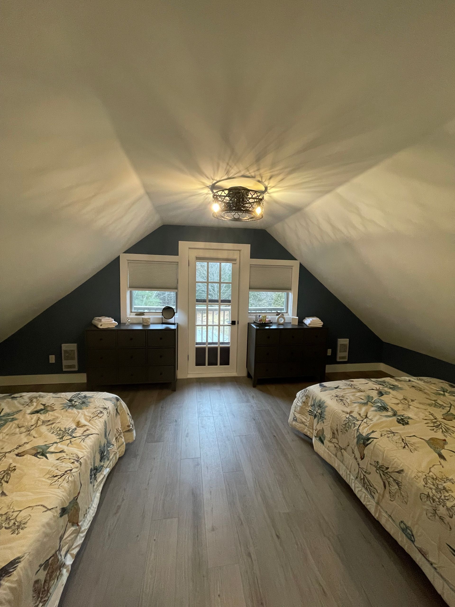 Bedroom in an attic with slanted ceiling. Two beds face each other, dark dresser between windows and a door.