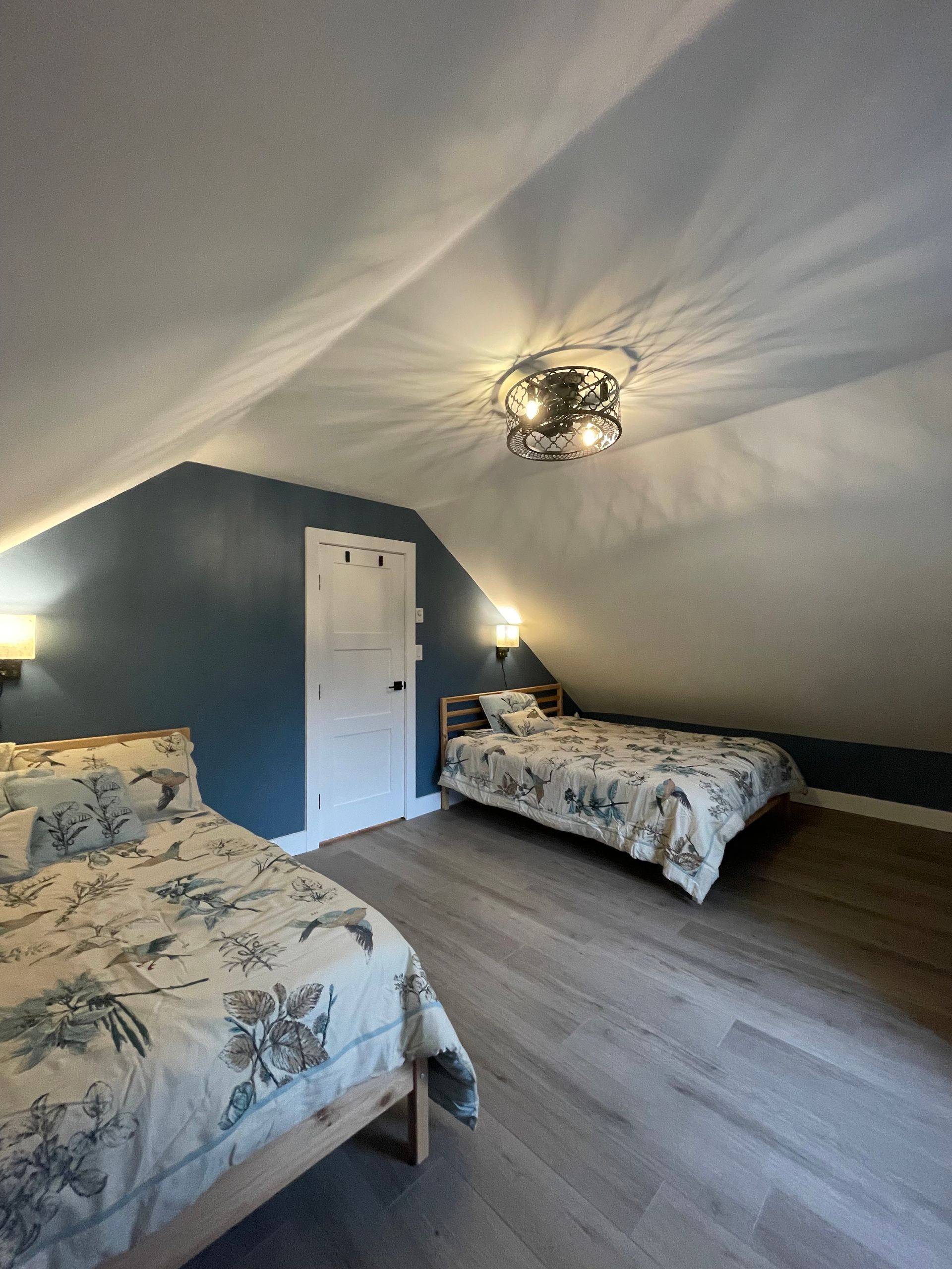 Bedroom with two beds under sloped ceiling, blue wall, wooden floors, and a decorative ceiling light.