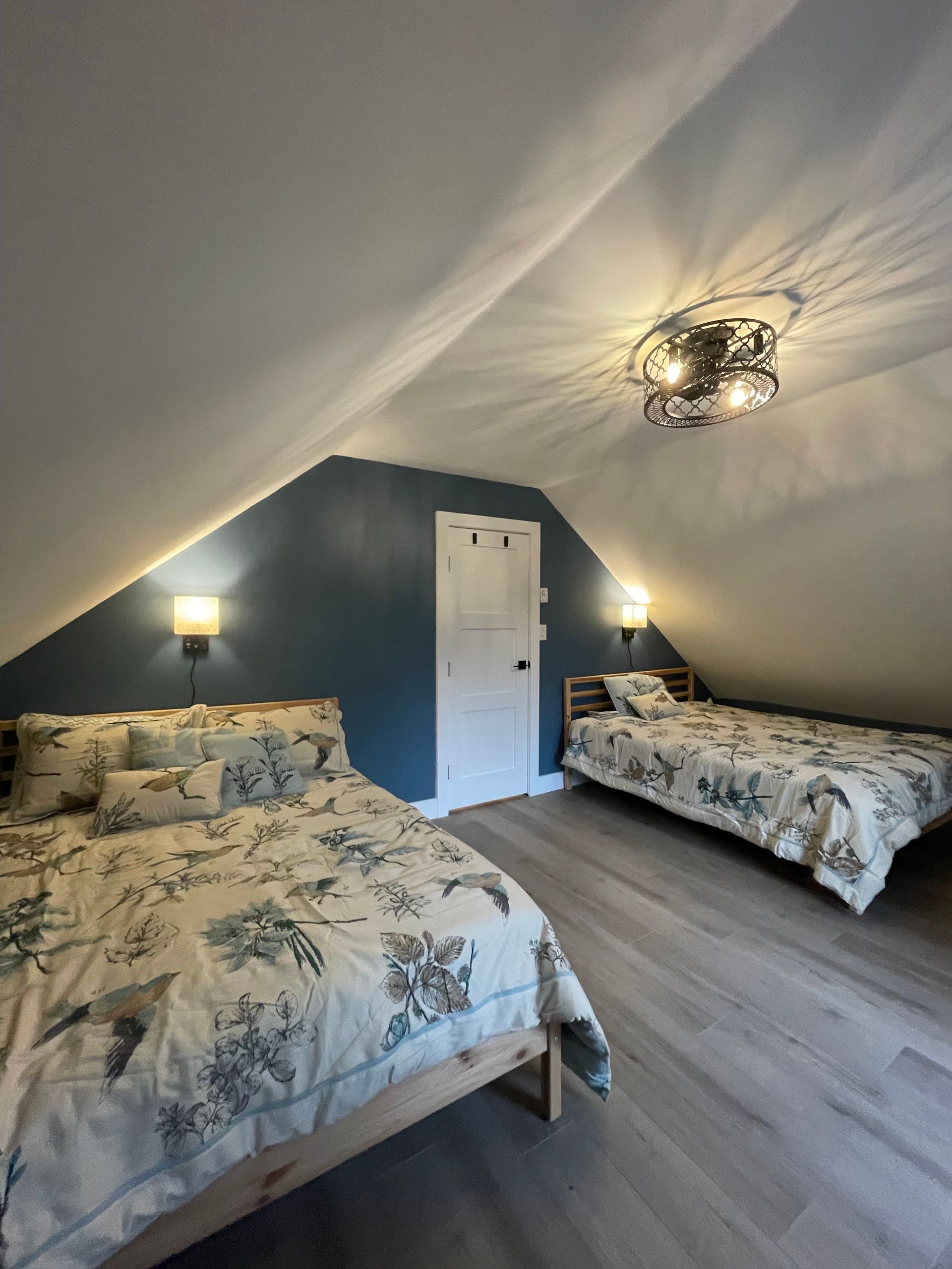 Bedroom with two beds under a sloped ceiling. Teal accent wall, wooden floors, sconces, and a decorative light fixture.