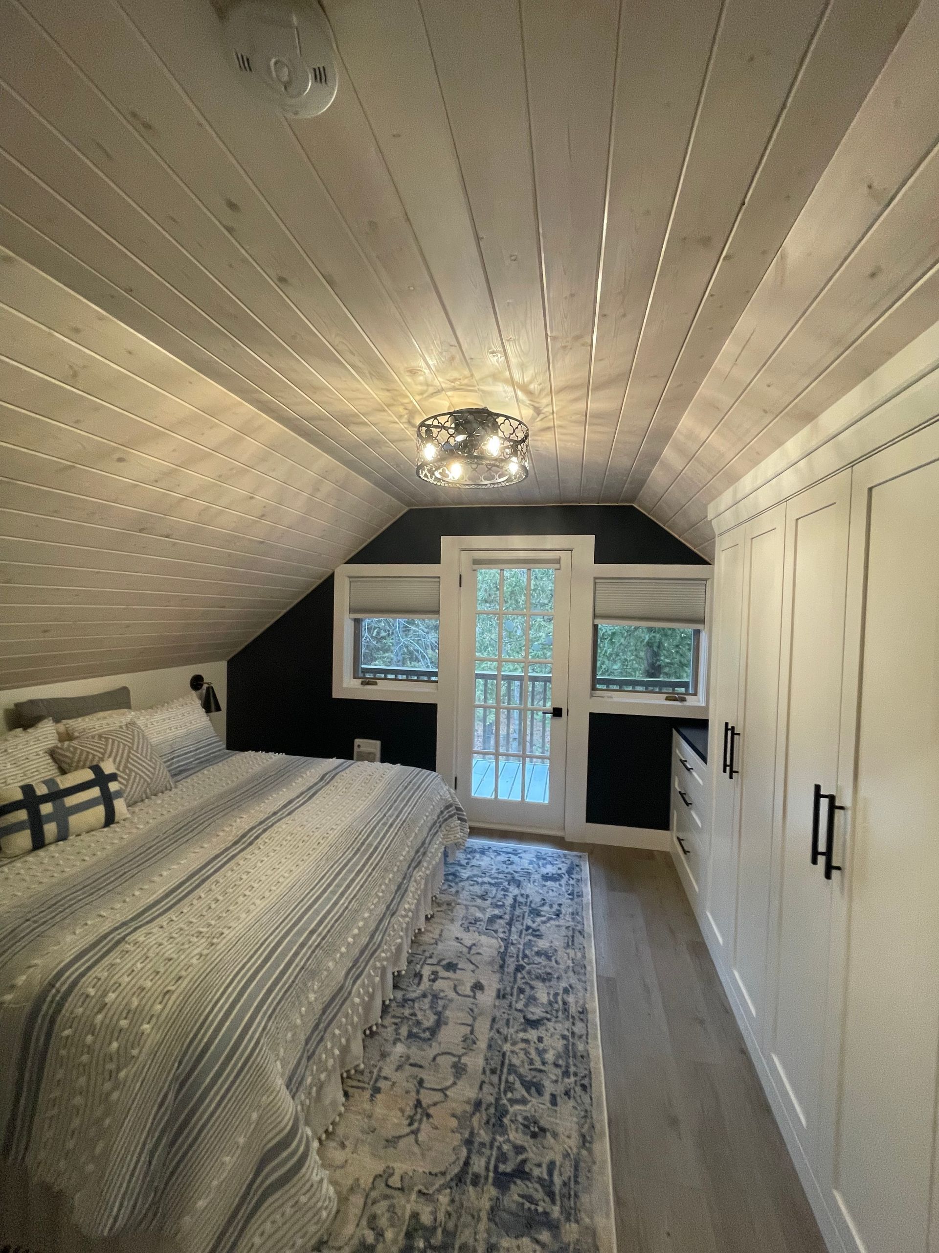 Bedroom with sloped ceiling, white wood paneling, bed with blue and white bedding, rug, door, and built-in white cabinets.