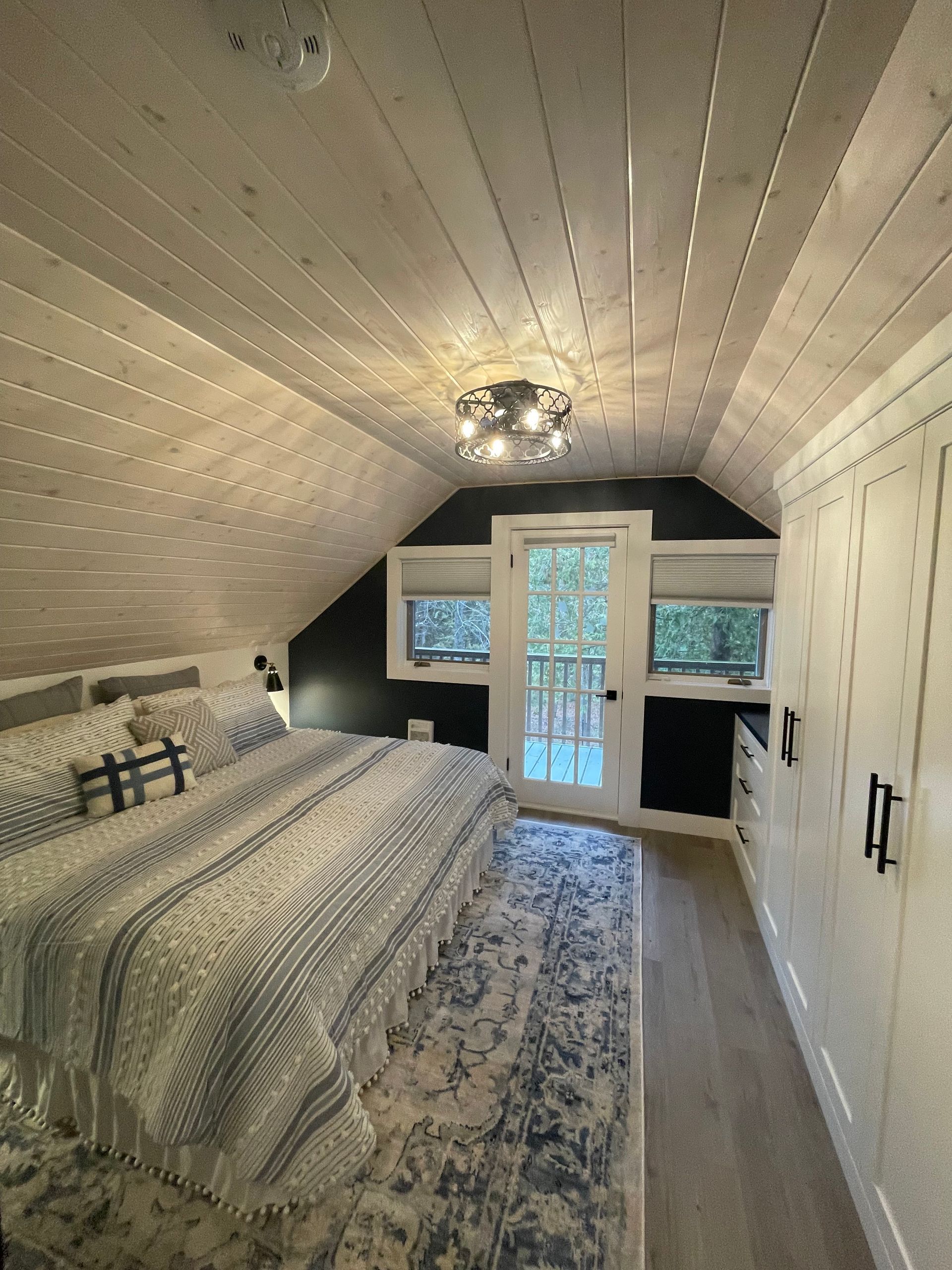 Bedroom with sloped ceiling, wood paneling, bed with blue bedding, closet, and door to balcony.