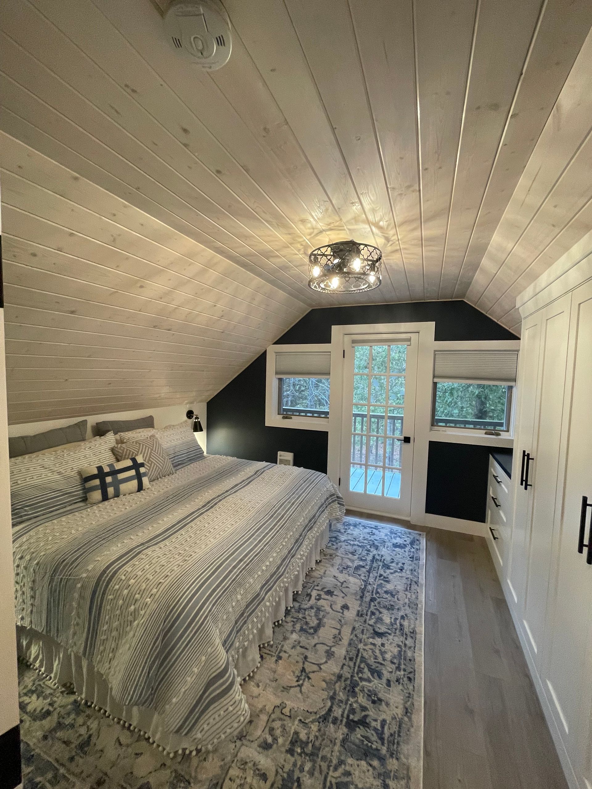 Bedroom with sloped ceiling, bed with blue bedding, rug, door to balcony, white walls, and dark accent wall.